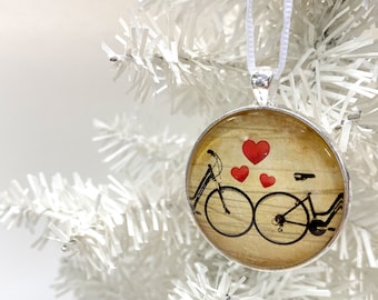 May include: A silver ornament with a brown background and a black silhouette of two bicycles with red hearts. The ornament is hanging from a white string on a white artificial Christmas tree.