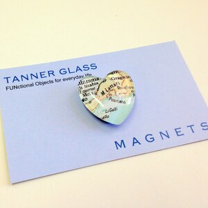 May include: A heart-shaped glass magnet featuring a map of the Amalfi Coast, Italy. The magnet is displayed on a light blue card with the text "TANNER GLASS FUNctional Objects for everyday life" and "MAGNETS".