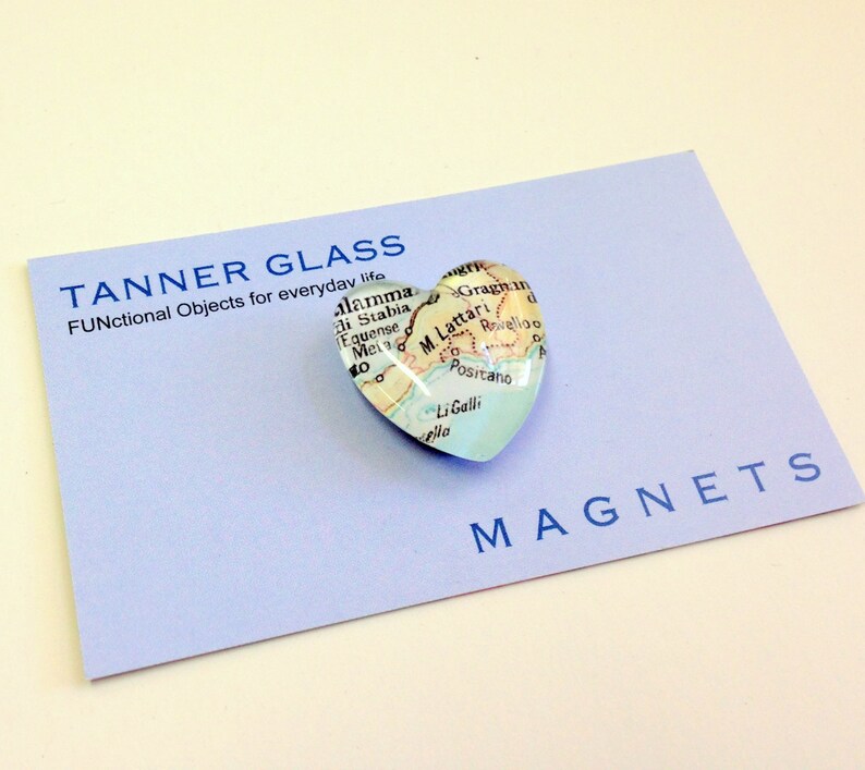 May include: A heart-shaped glass magnet featuring a map of the Amalfi Coast, Italy. The magnet is displayed on a light blue card with the text "TANNER GLASS FUNctional Objects for everyday life" and "MAGNETS".
