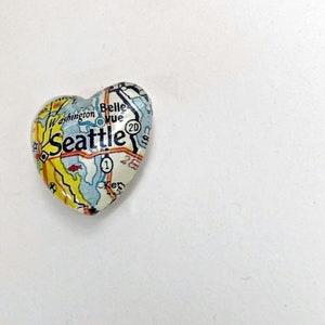 May include: A heart-shaped charm with a map of Seattle, Washington. The charm is made of glass and has a white background with blue and yellow details. The text "Seattle" is printed in black on the charm.