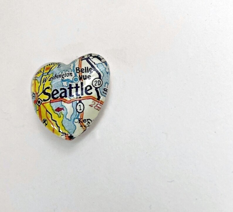 May include: A heart-shaped charm with a map of Seattle, Washington. The charm is made of glass and has a white background with blue and yellow details. The text "Seattle" is printed in black on the charm.