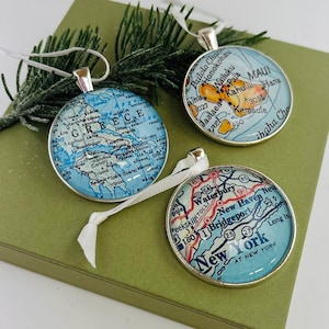 May include: Three round silver-toned ornaments featuring map prints under glass domes. One shows Greece, one shows Maui, and one shows New York. Each ornament has a white ribbon loop for hanging and is displayed with green pine needles.