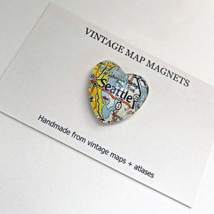 May include: A heart-shaped magnet with a vintage map of Seattle, Washington. The magnet is made from vintage maps and atlases. The text "VINTAGE MAP MAGNETS" and "Handmade from vintage maps + atlases" is printed on a white card.