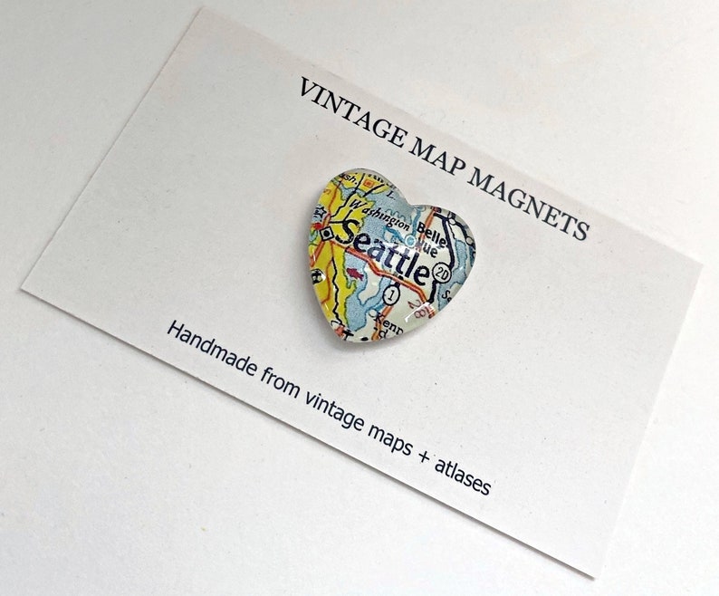 May include: A heart-shaped magnet with a vintage map of Seattle, Washington. The magnet is made from vintage maps and atlases. The text "VINTAGE MAP MAGNETS" and "Handmade from vintage maps + atlases" is printed on a white card.