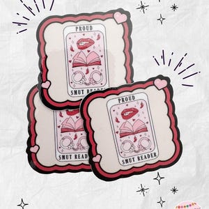 May include: Three stickers with a pink and red border. Each sticker has a white background with a graphic of an open book, handcuffs, and a pair of red lips. The word "PROUD" is at the top, and "SMUT READER" is at the bottom.