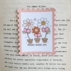 Peut inclure: Un autocollant rose en forme de timbre avec un motif floral. Trois fleurs souriantes dans des pots tiennent des livres roses, avec le texte "PROBABLY READING SMUT" en dessous. Le haut de l'autocollant indique "For Annie Wilkes".