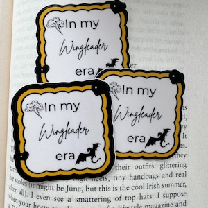 May include: Three stickers with a white background, black border, and yellow trim. The stickers have the text "In my Wingleader era" with a lightning bolt and dragon graphic. The stickers are on top of an open book.
