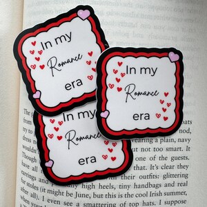 May include: Three stickers with the text "In my Romance era". Each sticker has a white background, a red border, and a black outline. Small red hearts and pink hearts decorate the stickers. The stickers are on top of an open book.
