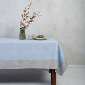 May include: A light blue tablecloth with a silver border, set on a wooden table. A vase with cotton flowers and a plate with a small snack are on the table.