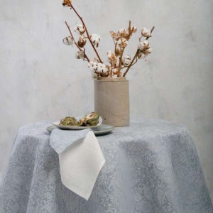 May include: A white linen tablecloth with a grey paisley pattern. A white napkin is folded on the table. A vase with cotton branches sits on the table.