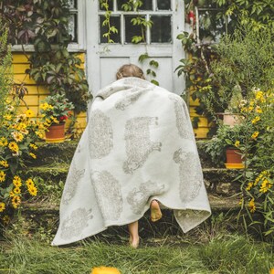 Può includere: Un bambino indossa una coperta bianca con un motivo di pecore grigie e scende una scalinata di pietra. I gradini sono circondati da erba verde e fiori gialli.