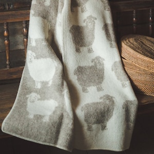 Può includere: Una morbida coperta di lana bianca e grigia con un motivo ripetitivo di pecore. La coperta è piegata e drappeggiata su una panca di legno.