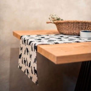 Może przedstawiać: Biały bieżnik stołowy z wzorem czarnych sylwet kotów. Bieżnik leży na drewnianym stole z wiklinowym koszem i białą miską.