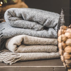 Op de afbeelding: Een stapel van drie grijze en beige dekens met franjes. De dekens zijn gevouwen en liggen op een donker houten oppervlak.