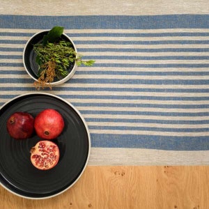 Op de afbeelding: Een zwart bord met een witte rand, met drie rode granaatappels, staat op een blauw-wit gestreept linnen tafelkleed.