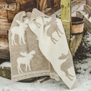 Könnte beinhalten: Eine weiche, beige und weiße Wolldecke mit einem Elch-Design. Die Decke ist gefaltet und über eine Holzoberfläche gelegt.