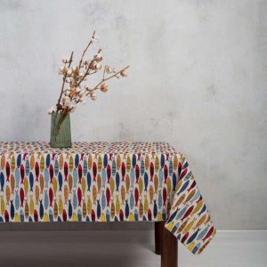 May include: A white tablecloth with a colorful fish pattern. The tablecloth is laid on a wooden table with a vase of cotton flowers on top.
