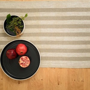 Puede incluir: Un plato negro con tres granadas rojas sobre un camino de mesa de lino beige a rayas.