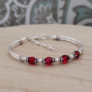 May include: A silver bangle bracelet with a delicate chain extender and a row of red crystal beads.