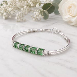 May include: A silver bracelet featuring a row of square, light green crystals. The bracelet has a delicate design with a toggle clasp and is accented with small, clear crystals. The background includes white flowers and greenery.