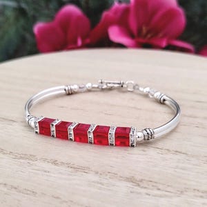 May include: A silver-toned bracelet featuring five square-cut, vibrant red crystal beads arranged in a row. Each red bead is separated by a small, clear crystal-encrusted spacer. The bracelet has a smooth, polished metal band and a toggle clasp.