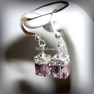 May include: A pair of silver dangle earrings with a small, square, purple crystal charm. The earrings are on a white display stand.