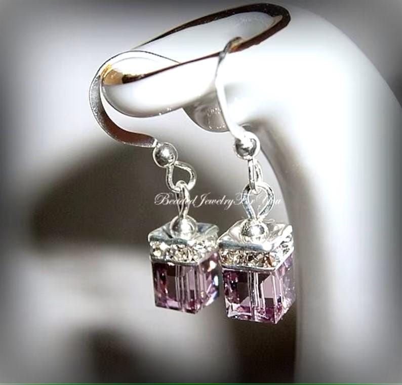 May include: A pair of silver dangle earrings with a small, square, purple crystal charm. The earrings are on a white display stand.