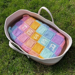 Op de afbeelding: Een witte geweven mand met een handvat, met daarin een pastelkleurig gebreid deken met de naam "TESSA" in wit.