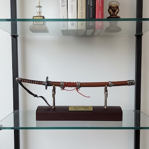 Pode incluir: Uma espada katana com um cabo de madeira castanho e uma lâmina prateada, exposta num suporte de madeira. O cabo é enrolado com cordão vermelho e preto. A espada está numa prateleira de vidro.
