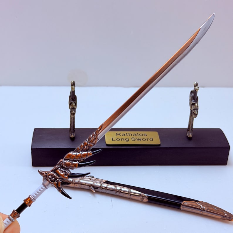 May include: A detailed, decorative long sword with a bronze and silver blade, resting on a wooden stand. The sword features intricate handle and guard designs. The text "Rathalos Long Sword" is on a gold plaque on the stand.