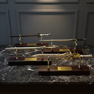 Può includere: Una collezione di spade decorative esposte su supporti in legno. Le spade presentano vari design, con lame dorate e argentate e impugnature ornate. Ogni spada è etichettata. L'esposizione è su una superficie di marmo.