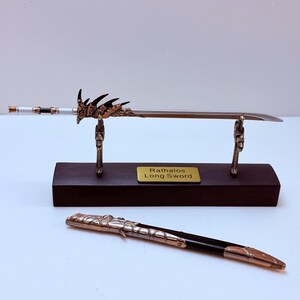 May include: A long sword with a silver blade and ornate bronze hilt, displayed on a wooden stand. The hilt features a dragon-like design. The scabbard is also visible. The text on the stand reads "Rathalos Long Sword."