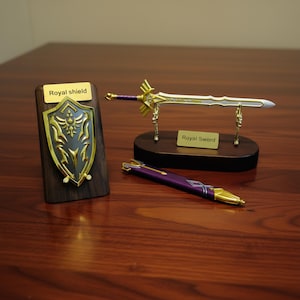 May include: A collection of miniature fantasy items, including a gold and silver sword on a wooden stand, a shield, and a purple pen. The shield and sword have gold accents and the text "Royal shield" and "Royal Sword".