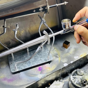 May include: A silver-colored sword is held in place with metal clamps during the painting process. An airbrush is used to apply paint to the sword. The sword is on a metal surface.
