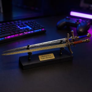 May include: A pair of miniature swords, one silver and one gold, mounted on a black rectangular stand. The blades feature intricate engravings, and the hilts are detailed with blue and brown accents. A gold plaque on the base reads Rellana's Twin Blades.