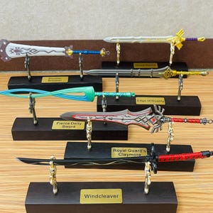Puede incluir: Colección de espadas en miniatura sobre soportes de exhibición. Las espadas varían en diseño, con colores como plata, oro, rojo, azul y verde. Cada espada está montada sobre una base de madera oscura con un soporte dorado. Etiquetas de texto identifican cada espada.