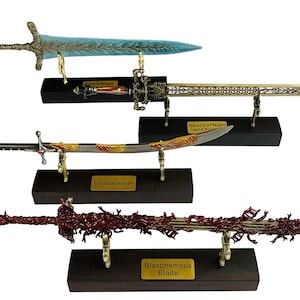 Puede incluir: Cuatro espadas decorativas en exhibición. Las espadas son de varios estilos, incluyendo una espada de hoja azul con un mango detallado, una espada dorada con patrones intrincados y una espada de color rojo. Cada espada está montada sobre una base negra con una placa de identificación.