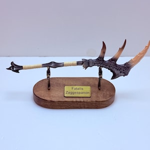 May include: A detailed replica of a weapon, the Fatalis Zaggespanon, with a wooden base. The weapon features a long, textured handle wrapped in beige material, a dark metal central section, and a multi-pronged head with brown and tan accents.
