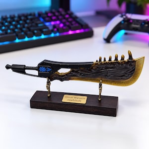 May include: A miniature Great Wyvern Jawblade replica sword featuring a black handle with blue accents and a textured, dark bronze-colored blade with a gold-toned edge. The sword is mounted on a rectangular dark wood base with a gold nameplate.