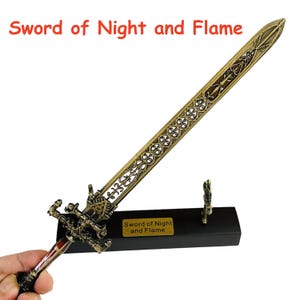 Op de afbeelding: Een sierlijk bronzen zwaard met een decoratief handvat en lemmet, rustend op een zwarte standaard. Het zwaard heeft ingewikkelde patronen en de tekst "Sword of Night and Flame" is zichtbaar.