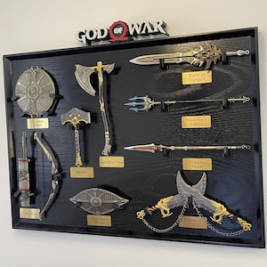 May include: A framed display featuring replicas of weapons from the video game God of War. Includes the Blade of Olympus, Leviathan Axe, Mjolnir, and Blades of Chaos. The display is mounted on a black wooden surface with gold nameplates.