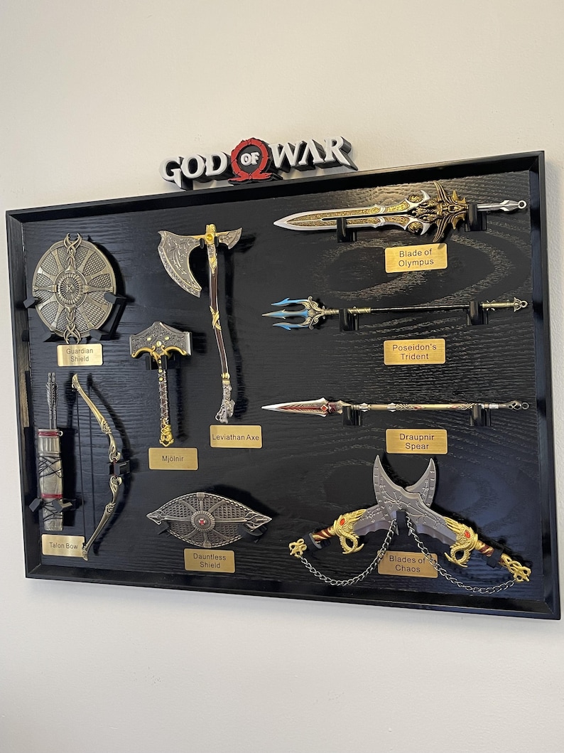 May include: A framed display featuring replicas of weapons from the video game God of War. Includes the Blade of Olympus, Leviathan Axe, Mjolnir, and Blades of Chaos. The display is mounted on a black wooden surface with gold nameplates.