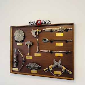 May include: A framed display of weapons from the "God of War" video game series. The collection includes a Guardian Shield, Leviathan Axe, Blade of Olympus, and Blades of Chaos. The display is mounted on a brown wooden board.