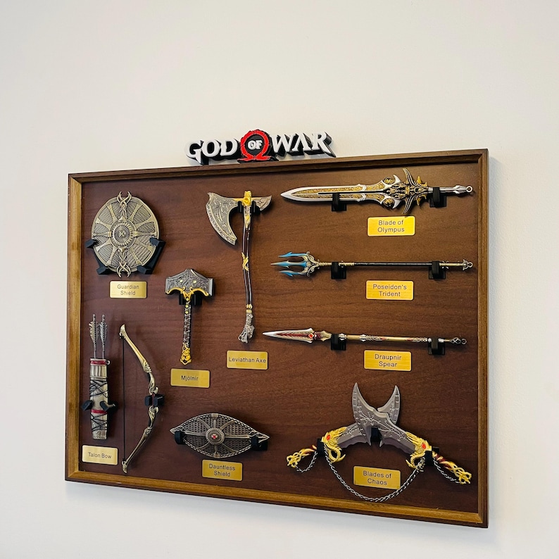 May include: A framed display of weapons from the "God of War" video game series. The collection includes a Guardian Shield, Leviathan Axe, Blade of Olympus, and Blades of Chaos. The display is mounted on a brown wooden board.