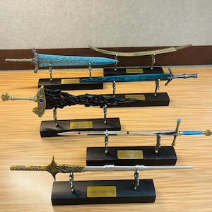 May include: A collection of detailed miniature swords, each displayed on a black stand. The swords vary in design, with colors including gold, silver, blue, and black. Each stand has a nameplate identifying the sword.
