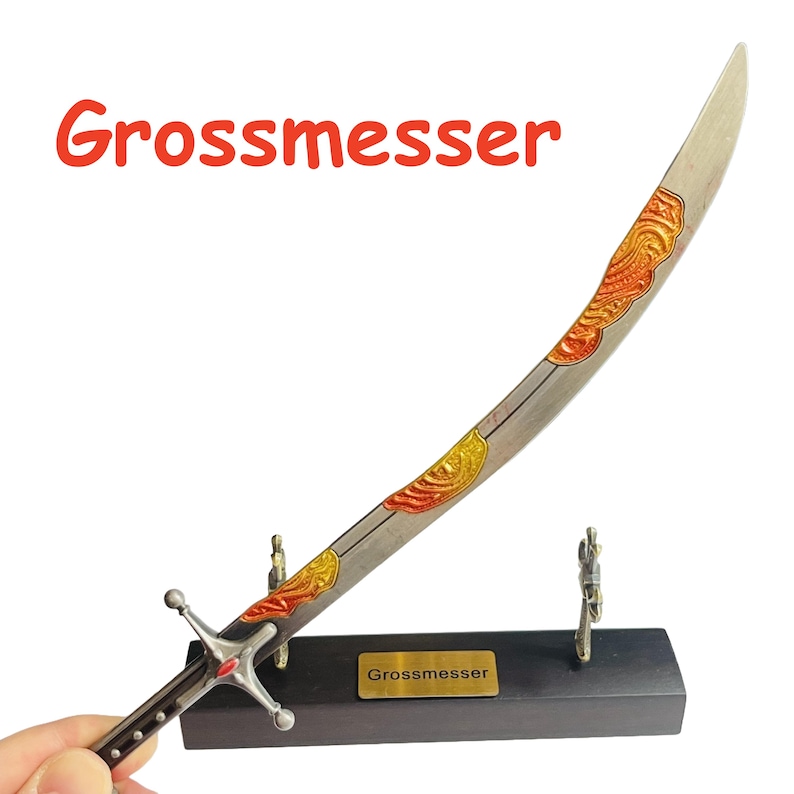 Op de afbeelding: Een decoratief zwaard met een gebogen lemmet, met sierlijke gouden en rode ontwerpen. Het zwaard is gemonteerd op een zwarte basis met het woord "Grossmesser" in het rood. De handgreep heeft een zilveren pareerstang en een rode edelsteen.