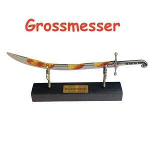Op de afbeelding: Een decoratief zwaard met een gebogen lemmet, met gouden en rode accenten. Het zwaard wordt tentoongesteld op een zwarte houten standaard met een gouden naamplaatje met de tekst "Grossmesser". Het woord "Grossmesser" staat ook in het rood boven het zwaard.