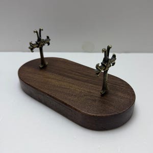 May include: A dark brown wooden display stand with two ornate metal crosses. The crosses are attached to the wooden base, which has an oval shape. The metal crosses have intricate details and a vintage appearance.