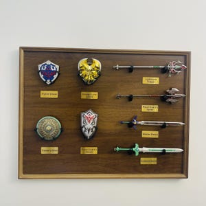 Pode incluir: Uma vitrine de madeira com réplicas de armas e escudos de um videogame. A vitrine inclui Hylian Shield, Savage Lynel Shield, Traveler's Shield, Royal Guard's Shield, Lightscale Trident, Royal Guard's Spear, Master Sword e Goddess Sword.