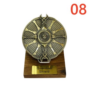 May include: A bronze-colored Guardian Shield, mounted on a wooden base. The shield features an intricate design with a central circular emblem and cross-like patterns. The number "08" is in the upper right corner. A gold plaque reads "Guardian Shield."